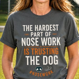 May include: A dark gray short-sleeve t-shirt featuring the white and orange text THE HARDEST PART OF NOSE WORK IS TRUSTING THE DOG with a small dog nose icon and the hashtag #NOSEWORK printed on the front chest area.