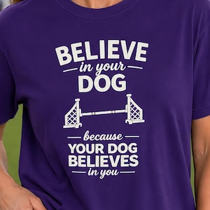 May include: A purple short-sleeved t-shirt featuring white text that reads BELIEVE in your DOG because YOUR DOG BELIEVES in you, with a graphic of a dog agility hurdle jump positioned between the two lines of text on the front of the shirt.