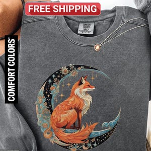 May include: A dark gray sweatshirt with a fox and moon graphic. The fox is orange and white, sitting on a crescent moon with floral and star details. The sweatshirt has the words "Comfort Colors" and "Pepper" printed on it.