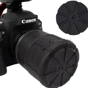 May include: Black Canon EOS camera lens with a black silicone lens cap. The lens cap has a starburst design with a small plus sign in the center.