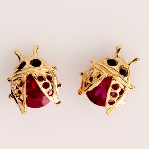 May include: A pair of gold ladybug earrings with red and black accents. The earrings feature a detailed design with a red gemstone in the center.