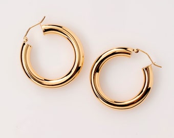 May include: A pair of gold-coloured hoop earrings. The earrings are thick and round, with a polished, reflective surface. The hoops have a classic design and are displayed on a white background.