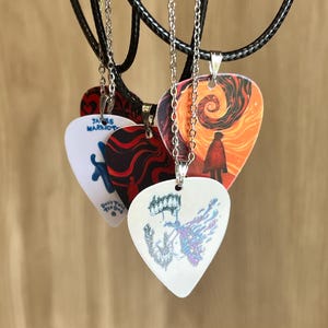 May include: A collection of four guitar pick pendants hanging from black cords and silver chains. The picks feature various graphic designs, including a figure in a swirling orange landscape, a white pick with blue text reading James Marriott, and a white pick with a colourful abstract illustration.