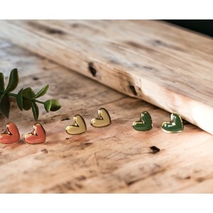 May include: Three pairs of heart-shaped earrings in different colors: yellow, coral, and green. The earrings are gold-toned and have a glossy finish.