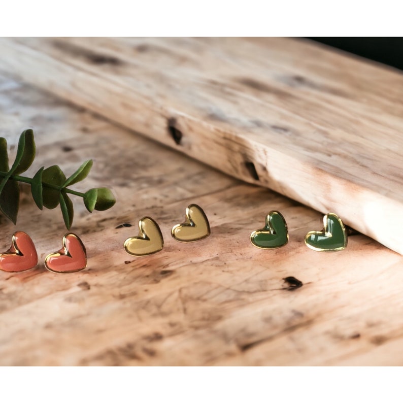 May include: Three pairs of heart-shaped earrings in different colors: yellow, coral, and green. The earrings are gold-toned and have a glossy finish.