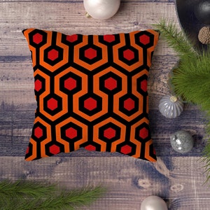 Op de afbeelding: Een vierkant kussen met een geometrisch patroon van oranje en rode zeshoeken op een zwarte achtergrond. Het kussen ligt op een houten oppervlak met witte en zilveren ornamenten.