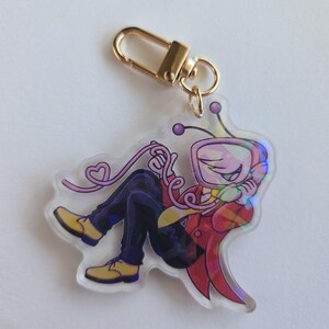 May include: A clear acrylic keychain featuring a cartoon character with a TV-shaped head, wearing a red jacket, yellow shirt, and blue pants. The keychain has a gold-colored clasp and a pink heart-shaped detail.