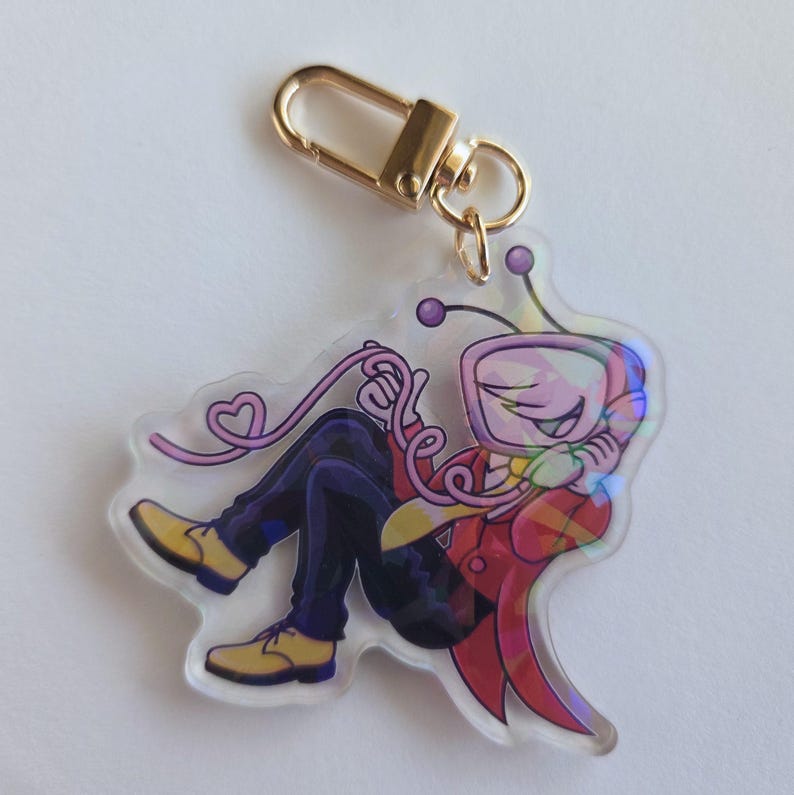 May include: A clear acrylic keychain featuring a cartoon character with a TV-shaped head, wearing a red jacket, yellow shirt, and blue pants. The keychain has a gold-colored clasp and a pink heart-shaped detail.