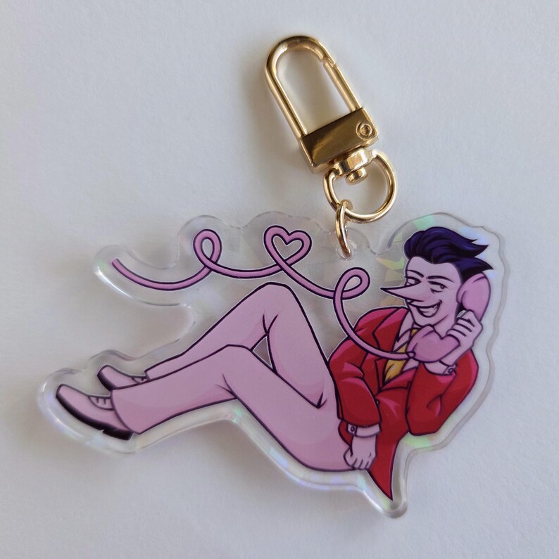 May include: A clear acrylic keychain featuring a cartoon character in a red suit and pink pants, holding a pink phone. The phone cord is pink and shaped like a heart. A gold-colored clasp is attached.