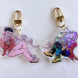 May include: Two acrylic keychains with gold-colored clasps. One features a character in a red suit and pink pants, holding a phone. The other keychain has a character in a colorful outfit. Both have a pink phone cord with hearts.