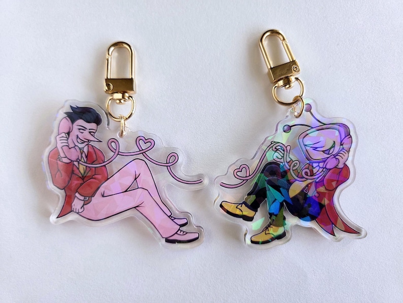 May include: Two acrylic keychains with gold-colored clasps. One features a character in a red suit and pink pants, holding a phone. The other keychain has a character in a colorful outfit. Both have a pink phone cord with hearts.