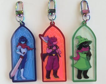 May include: Three character keychains with iridescent clasps. Each keychain features a different character from a video game, set against a stained-glass window design. The characters are in shades of blue, red, and green, with detailed artwork and vibrant colors.