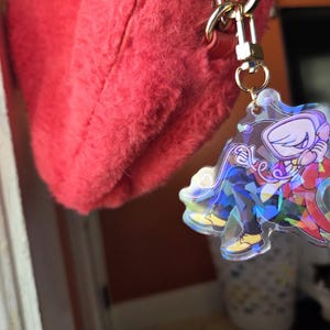 May include: A clear acrylic keychain featuring a cartoon character in a red suit and yellow shoes. The keychain has a gold-colored chain and clasp, attached to a fluffy red bag. The character is holding a phone.