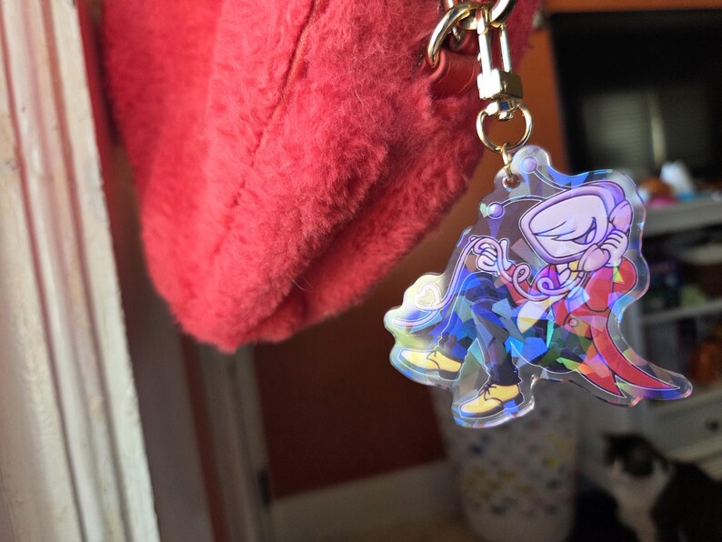May include: A clear acrylic keychain featuring a cartoon character in a red suit and yellow shoes. The keychain has a gold-colored chain and clasp, attached to a fluffy red bag. The character is holding a phone.