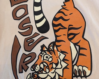 May include: A light pink t-shirt featuring a cartoon tiger in orange and black stripes, with the word "LOVER" in brown letters. The tiger is in a crouching pose, with its tail curved. The design has a playful, vintage aesthetic.