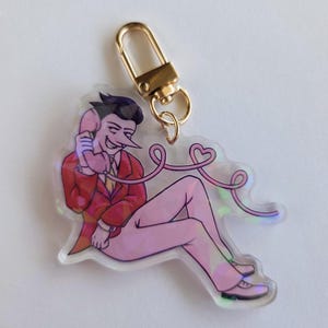May include: A clear acrylic charm featuring a cartoon character in a red suit and pink skin, holding a pink telephone. The charm has a gold-colored clasp and a pink cord ending in a heart shape.