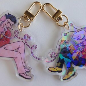 May include: Two acrylic keychains with gold-colored clasps. One keychain features a pink figure in a red suit holding a phone. The other keychain has a colorful, holographic figure holding a phone. Both have heart-shaped phone cords.