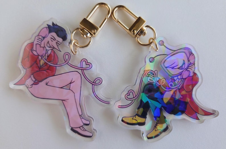 May include: Two acrylic keychains with gold-colored clasps. One keychain features a pink figure in a red suit holding a phone. The other keychain has a colorful, holographic figure holding a phone. Both have heart-shaped phone cords.