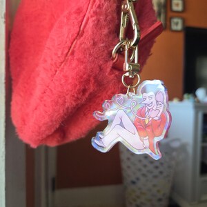 May include: A red fluffy handbag with a gold-colored clasp and a charm. The charm features a cartoon illustration of a person in a red jacket and white pants, holding a phone. The charm has a holographic finish.