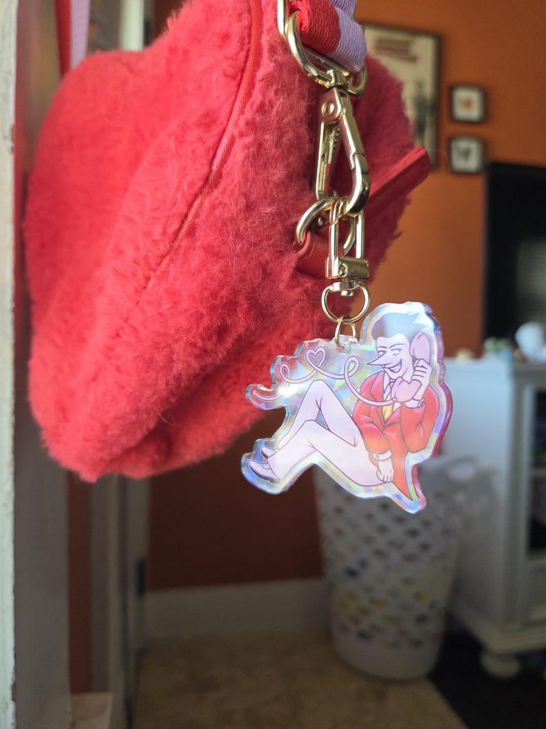 May include: A red fluffy handbag with a gold-colored clasp and a charm. The charm features a cartoon illustration of a person in a red jacket and white pants, holding a phone. The charm has a holographic finish.