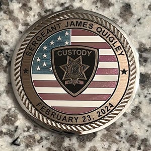 May include: A commemorative coin with a detailed design. The coin features the American flag, the text "SERGEANT JAMES QUIGLEY", and "FEBRUARY 23, 2024". A badge with the word "CUSTODY" is in the center. The coin has a silver and gold finish.