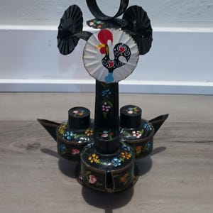 May include: A black metal decorative oil lamp with three oil containers and a central support featuring a rooster design. The containers and support are adorned with colorful floral patterns. The lamp has a handle and two fan-shaped elements.