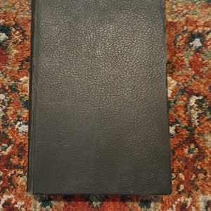 May include: A closed, dark gray book with a textured cover. The book is rectangular and appears to be leather-bound. The cover has a subtle, embossed pattern. The book is lying on a patterned rug with shades of red, orange, and brown.