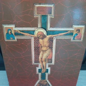 Puede incluir: Un icono religioso de madera que representa la crucifixi&oacute;n de Jesucristo. La cruz es de color turquesa con un halo dorado alrededor de la cabeza de Jes&uacute;s. El fondo es de color marr&oacute;n rojizo con un patr&oacute;n agrietado. Dos im&aacute;genes m&aacute;s peque&ntilde;as est&aacute;n a cada lado de la cruz.