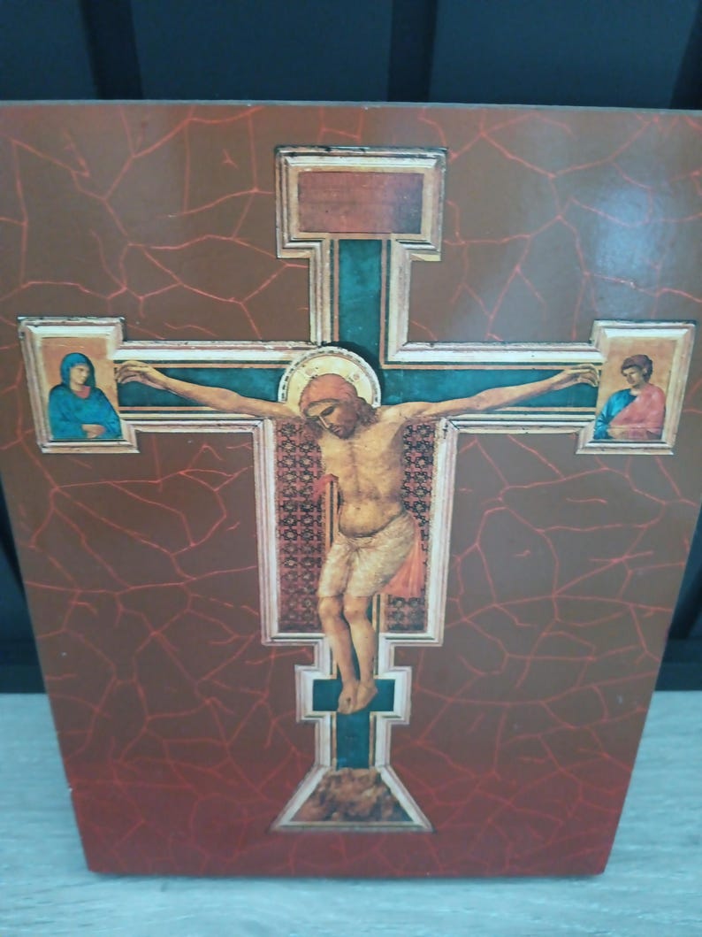 Puede incluir: Un icono religioso de madera que representa la crucifixi&oacute;n de Jesucristo. La cruz es de color turquesa con un halo dorado alrededor de la cabeza de Jes&uacute;s. El fondo es de color marr&oacute;n rojizo con un patr&oacute;n agrietado. Dos im&aacute;genes m&aacute;s peque&ntilde;as est&aacute;n a cada lado de la cruz.