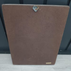 Puede incluir: Una placa rectangular de madera marr&oacute;n con un colgador de metal en la parte superior. La placa tiene una forma ligeramente trapezoidal y una superficie lisa. El fondo es una pared gris oscuro y una superficie de madera gris claro.