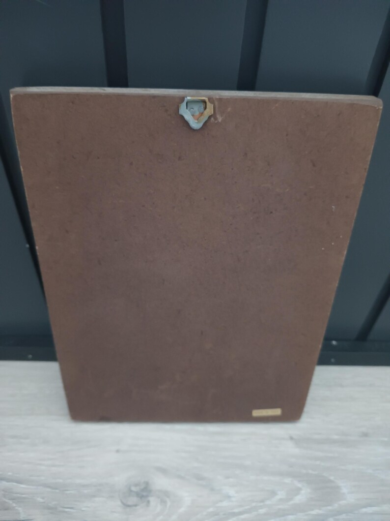 Puede incluir: Una placa rectangular de madera marr&oacute;n con un colgador de metal en la parte superior. La placa tiene una forma ligeramente trapezoidal y una superficie lisa. El fondo es una pared gris oscuro y una superficie de madera gris claro.