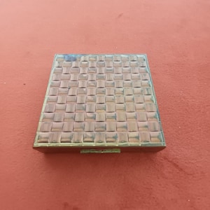 May include: A square, vintage compact case with a woven pattern on the lid. The case is made of a metallic material with a bronze and copper color. The background is a solid, reddish-orange color.