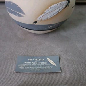 May include: A cream-colored pottery bowl with a gray rim and a gray band. The bowl is decorated with gray and white feather designs. Includes a tag that says "GREY FEATHER" by Desert Pueblo Pottery.