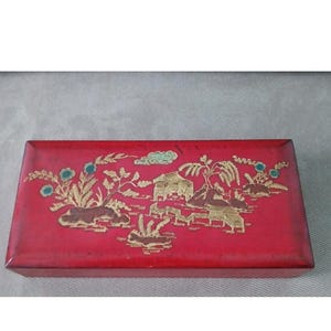 May include: A rectangular red box with a gold and teal landscape design. The box features a detailed illustration of buildings, trees, and clouds. The box is likely made of wood and is a decorative item.
