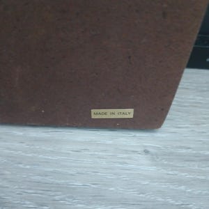 Puede incluir: Un objeto rectangular marr&oacute;n con una superficie texturizada. Una peque&ntilde;a etiqueta rectangular con las palabras "MADE IN ITALY" est&aacute; adherida. El objeto est&aacute; sobre una superficie de madera gris claro.