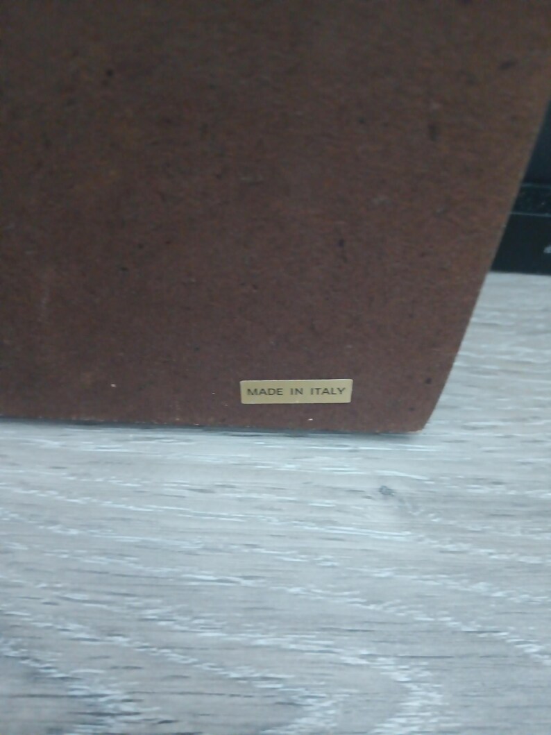 Puede incluir: Un objeto rectangular marr&oacute;n con una superficie texturizada. Una peque&ntilde;a etiqueta rectangular con las palabras "MADE IN ITALY" est&aacute; adherida. El objeto est&aacute; sobre una superficie de madera gris claro.