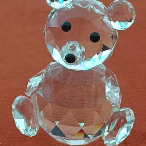 May include: A clear crystal bear figurine with a faceted design. The bear has black eyes and a black nose. The ears, head, body, arms, and legs are all made of clear crystal. The bear is sitting on a red-orange surface.