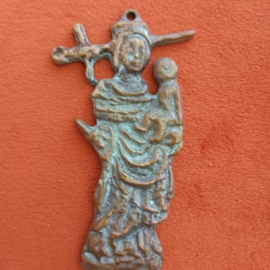 May include: A bronze-colored pendant depicting a figure holding a child, with a cross-like element above the head. The pendant has a loop at the top for hanging and shows intricate details in the clothing and facial features. The background is a solid orange color.