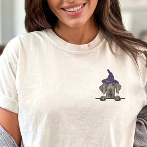 May include: Cream-colored t-shirt featuring a small graphic of a gray dog wearing a purple witch hat. The dog is peeking over a black line. The shirt has short sleeves and a relaxed fit, suitable for casual wear.