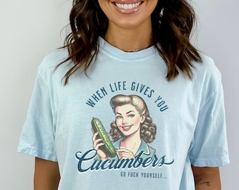 May include: Light blue t-shirt with a vintage-style graphic. The graphic features a woman holding a cucumber, with the text "WHEN LIFE GIVES YOU CUCUMBERS GO FUCK YOURSELF..." printed in a retro font.