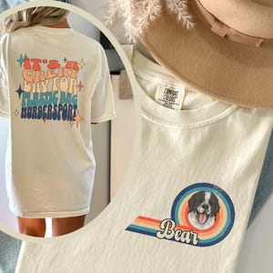 May include: Cream-colored t-shirt with a graphic of a dog and the word "Bear". The back of the shirt has the text "It's a great day for plastic bag murdersport" in retro colors. The shirt is a Comfort Colors brand.