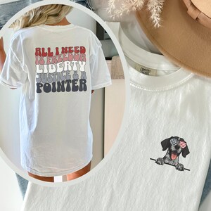 May include: White t-shirt with a graphic print on the back that says "All I need is freedom, liberty, justice & a pointer". The front of the shirt features a black and white dog wearing sunglasses with an American flag design.