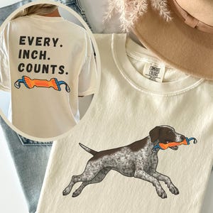 May include: Cream-colored t-shirt featuring a brown and white dog running with an orange and blue toy. The shirt also has the text "EVERY. INCH. COUNTS." on the back. The shirt is made of a soft material.