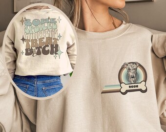 May include: A beige sweatshirt with a graphic print on the front. The graphic features a dog's head with a rainbow behind it and the text "Rosie". The back of the sweatshirt has a graphic print that says "Some Spoiled Whippets Treat Bitch" with stars.
