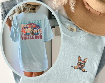 May include: Light blue t-shirt with a graphic of a cartoon cattle dog wearing sunglasses and a palm tree design. The text "Life is better with a cattle dog" is printed on the back of the shirt.