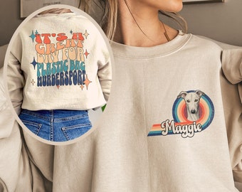 May include: A beige sweatshirt with a rainbow graphic and the text "Maggie" on the front. The graphic features a white dog with brown eyes. The back of the sweatshirt has a retro-style graphic with the text "It's a great day for plastic bag murder sport!"