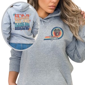 May include: A grey hooded sweatshirt with a rainbow design and a dog's face in a circle. The text "Gabe" is below the dog's face. The back of the sweatshirt has the text "It's a Great Day for Plastic Bag Murder-sport" in a retro font.