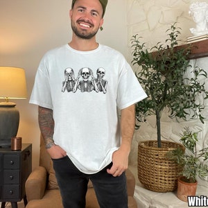 K&ouml;nnte beinhalten: Wei&szlig;es T-Shirt mit einer schwarz-wei&szlig;en Grafik von drei Skeletten in der "Nichts sehen, nichts h&ouml;ren, nichts sagen"-Pose. Das T-Shirt hat eine lockere Passform.