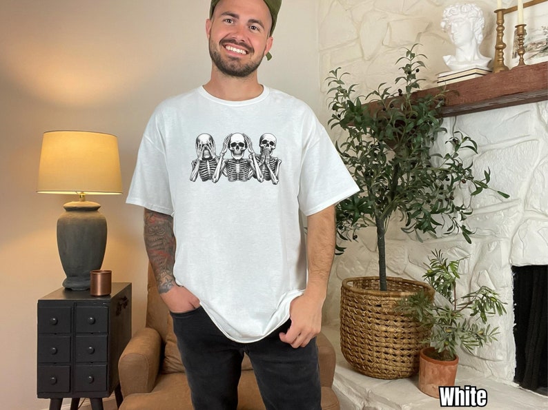 K&ouml;nnte beinhalten: Wei&szlig;es T-Shirt mit einer schwarz-wei&szlig;en Grafik von drei Skeletten in der "Nichts sehen, nichts h&ouml;ren, nichts sagen"-Pose. Das T-Shirt hat eine lockere Passform.