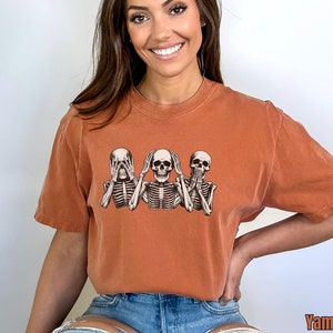 K&ouml;nnte beinhalten: Gebrannt orangefarbenes T-Shirt mit einer Grafik von drei Skeletten in der Pose &bdquo;nichts sehen, nichts h&ouml;ren, nichts sagen&ldquo;. Das Shirt hat eine lockere Passform und kurze &Auml;rmel. Das Wort &bdquo;Yam&ldquo; ist unten rechts aufgedruckt.